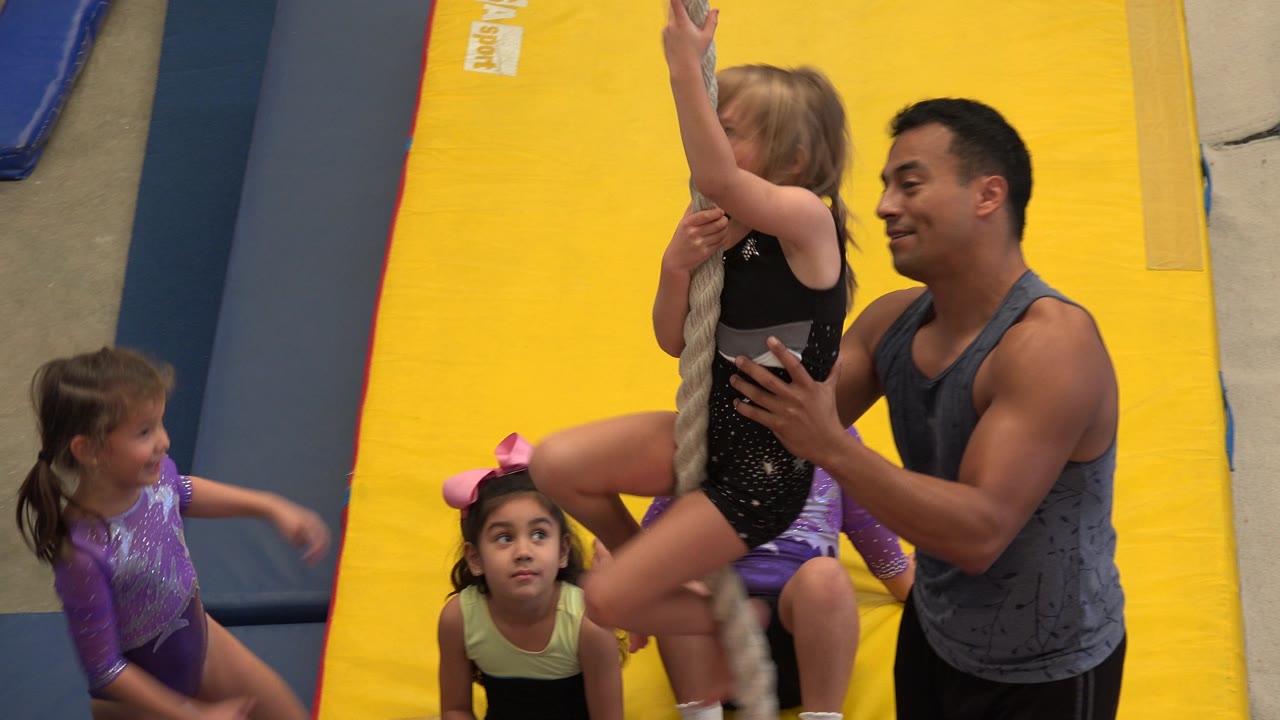 In Cabo San Lucas, Mexico, the girl remains on the rope with one arm raised as the coach holds her at the waist and two seated children look toward the activity.