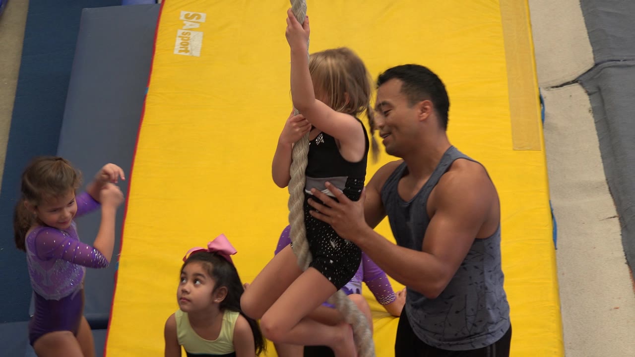 Coach holds a girl on a rope while children sit on mats.