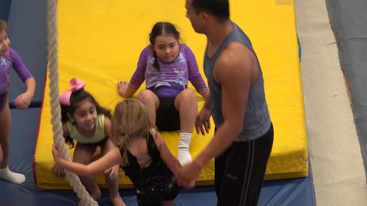 Children and an adult coach beside a yellow gymnastics mat and hanging rope.