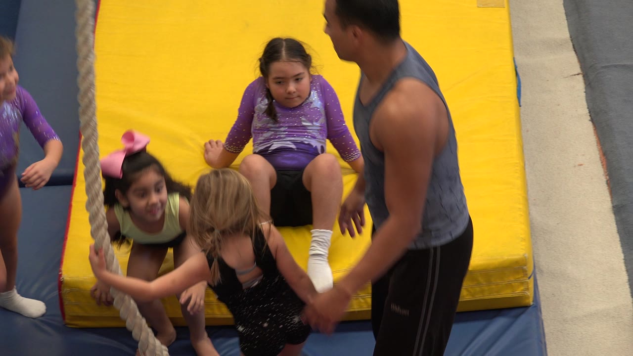 Several children remain close to the hanging rope while the adult coach faces them beside the yellow mat in the Cabo San Lucas gymnastics space.