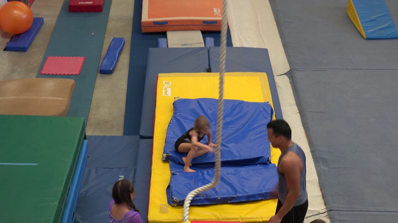 Young girls and an adult instructor near a hanging rope in a padded gymnastics area.
