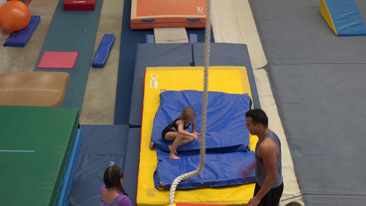 A child remains crouched on the blue mat beneath the rope while the adult instructor stands to the side and other children wait at the edge of the gymnastics area in Cabo San Lucas, Mexico.