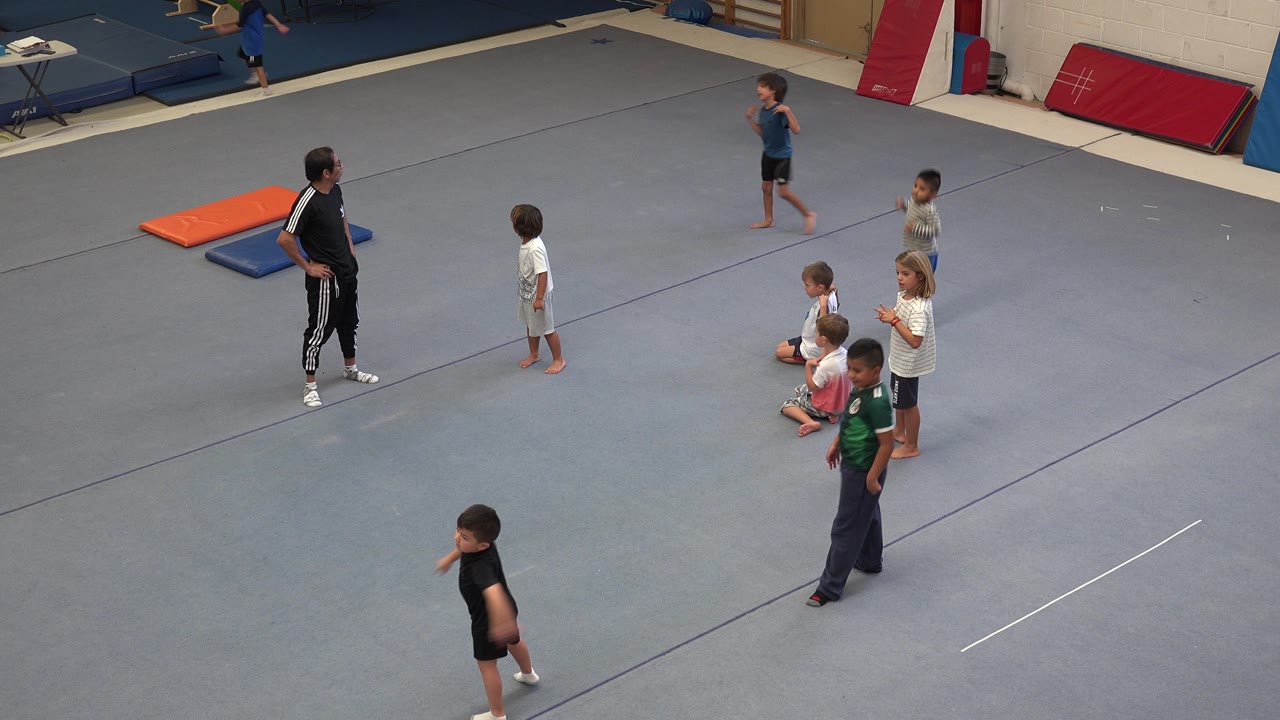 The coach watches from the left as children stand or walk across the gray court, with orange, blue, and red gym mats along the back wall in Cabo San Lucas, Mexico.