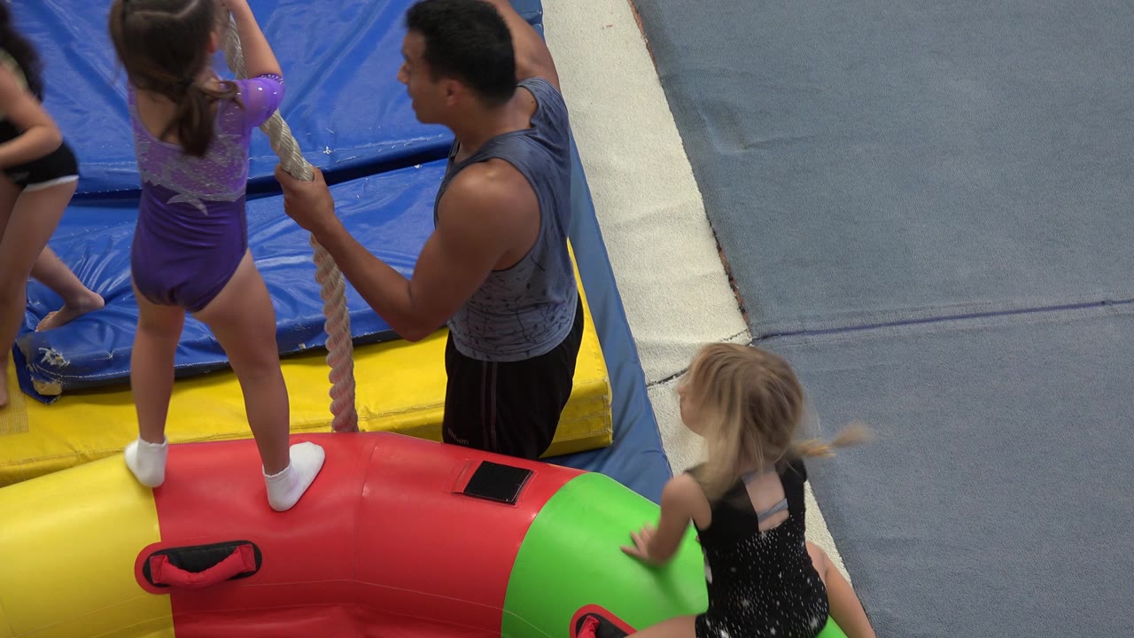 Child in purple leotard holding a rope beside a coach and padded mats.