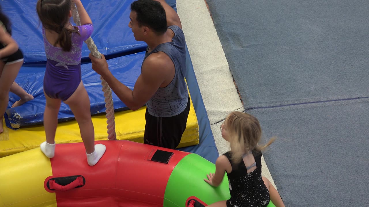 In Cabo San Lucas, the child remains on the padded obstacle with both hands near the rope while the coach stands close and another child approaches the green end of the pad.
