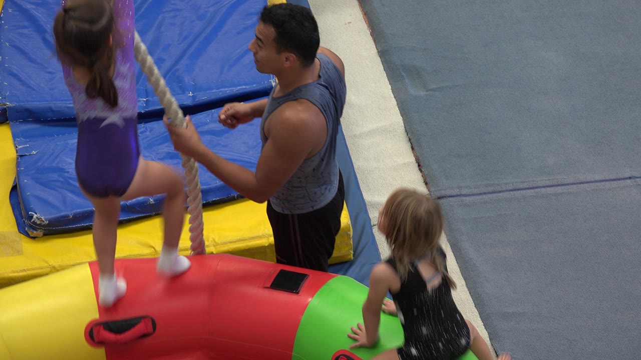 Child lifting on a rope from a padded obstacle as a coach stands nearby.