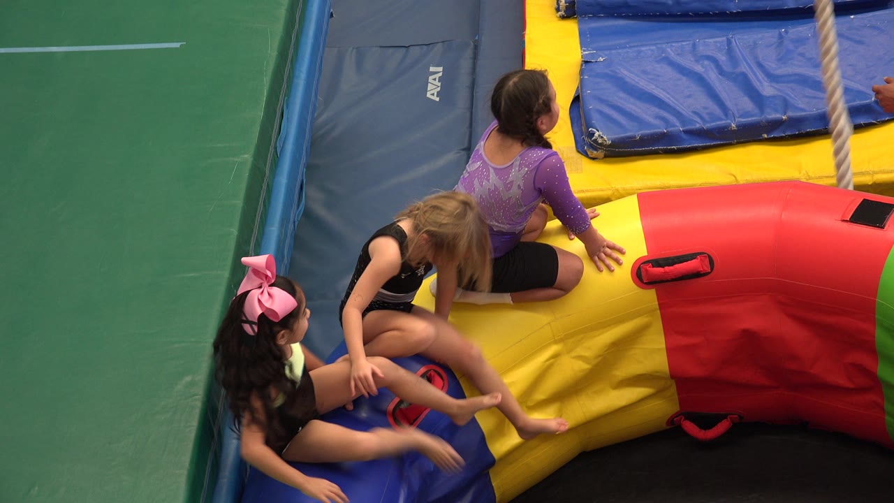Three girls sit and climb on a colorful inflatable inside a padded gym area.