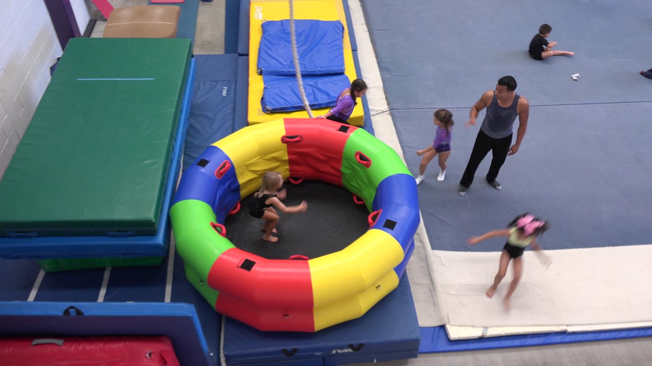 High-angle frame from Cabo San Lucas shows several children and an adult coach near blue mats, a white tumbling strip, and a multicolor inflatable trampoline ring.