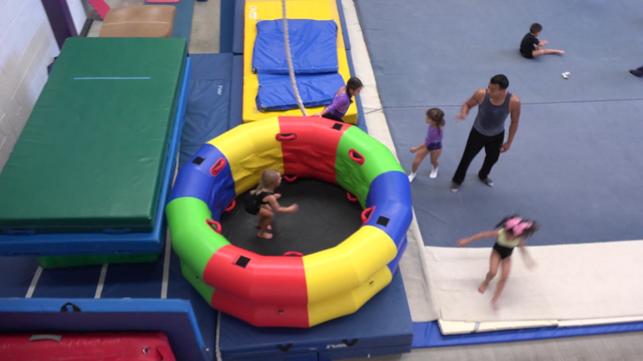 A child in motion crosses the white tumbling strip while other children and an adult coach stand near the multicolor inflatable ring in the Cabo San Lucas gym.