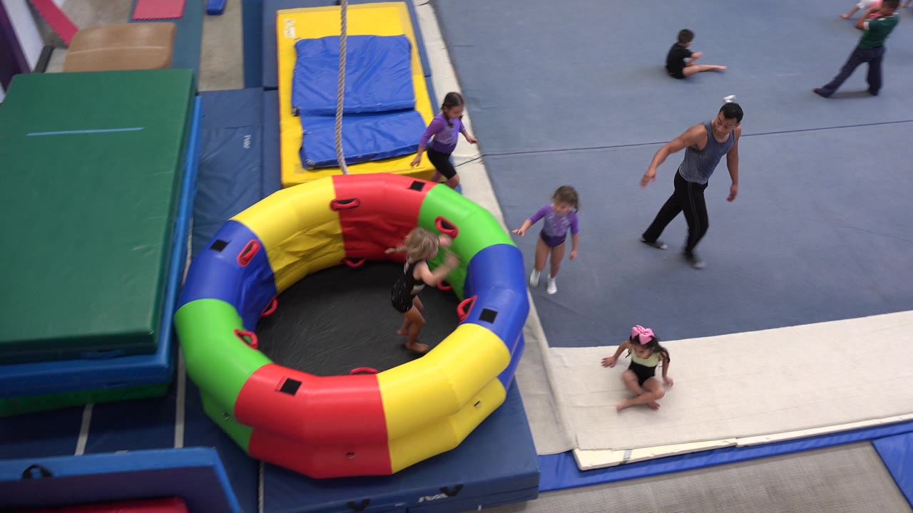 Children gather near an inflatable ring while a coach walks across the blue gym floor.
