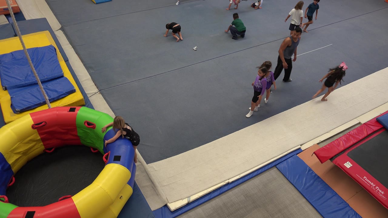 Children running on a grey gym floor near mats and trampolines.