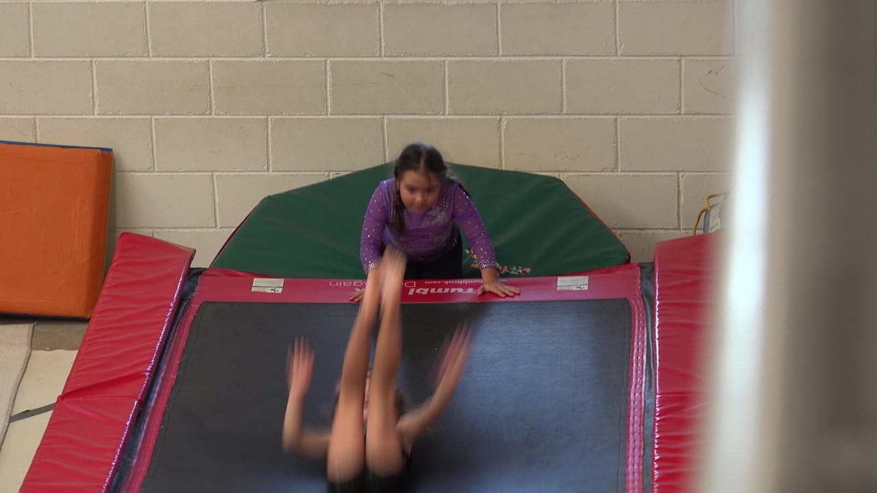 Child upside down on a trampoline mat while another child waits at the far edge.