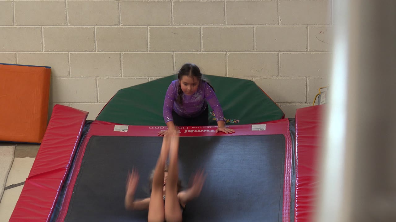 In Cabo San Lucas, the flipping child has legs lifted near the center of the black mat while the child in purple stays braced at the far padded edge.