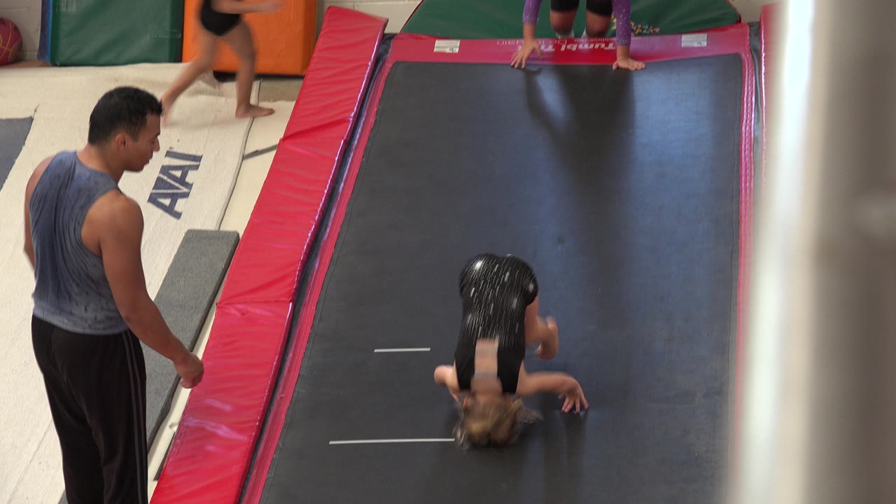 Girl inverted on a black gymnastics mat beside an adult instructor.