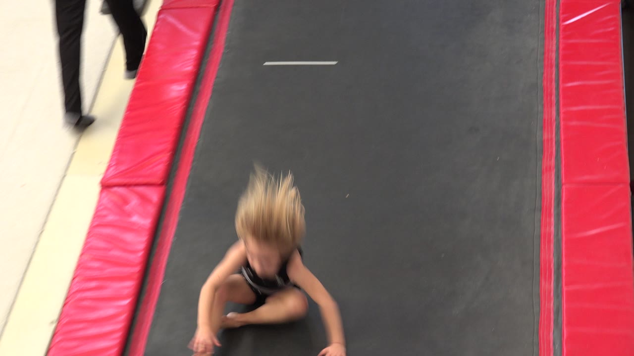 Young girl landing low on a black trampoline bordered by red pads.