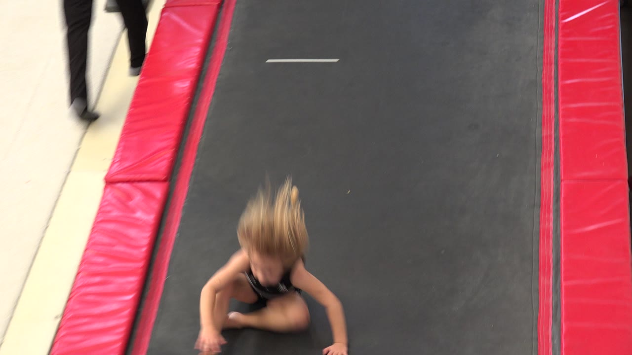The child gymnast is crouched near the lower edge of the black trampoline in Cabo San Lucas, with red side padding and beige flooring visible around the lane.