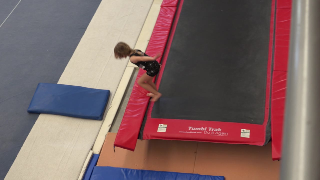 In Cabo San Lucas, Mexico, a young girl in a black leotard stands beside a red-edged trampoline with a blue mat on the gray gym floor.