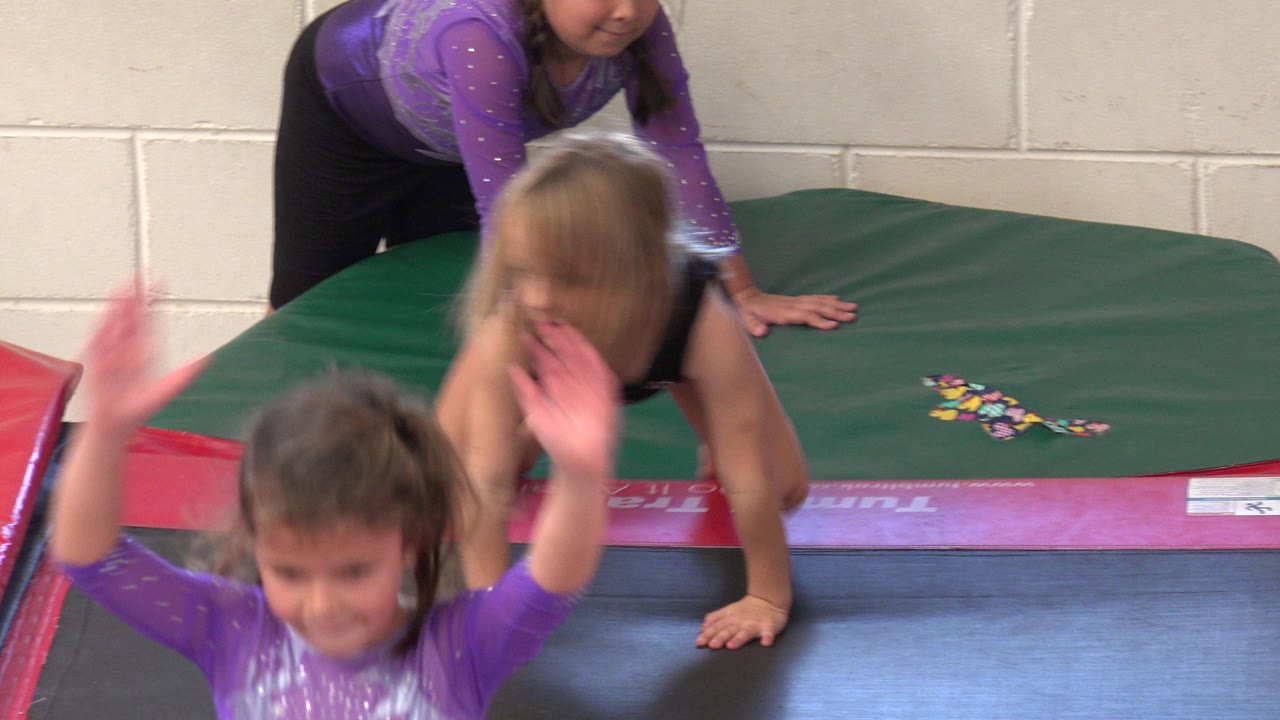 Children and instructor around a trampoline mat in a gym space.
