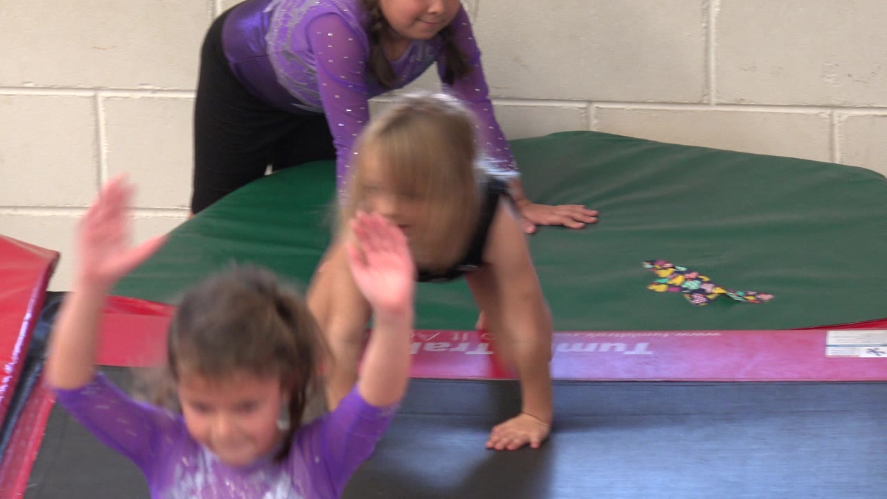 At 0:01 in Cabo San Lucas, Mexico, a child in purple passes close to the camera while the girl in black leans forward on the trampoline edge and the instructor stays behind her.