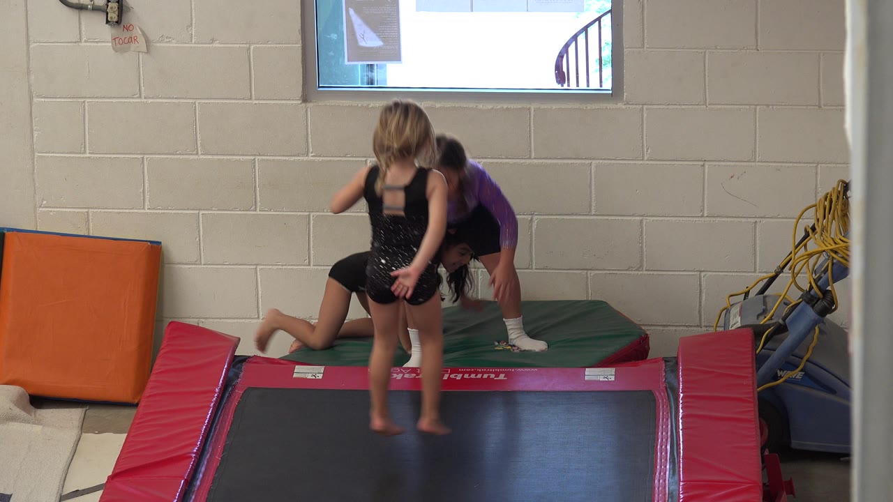 Three children play around a rectangular trampoline in Cabo San Lucas, Mexico, with padded mats, a block wall, and a high window behind them.