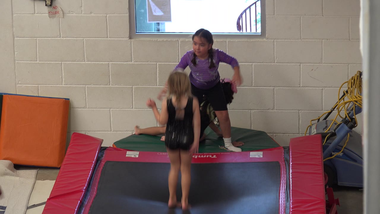 A child jumps on the trampoline while another stands on the mat.