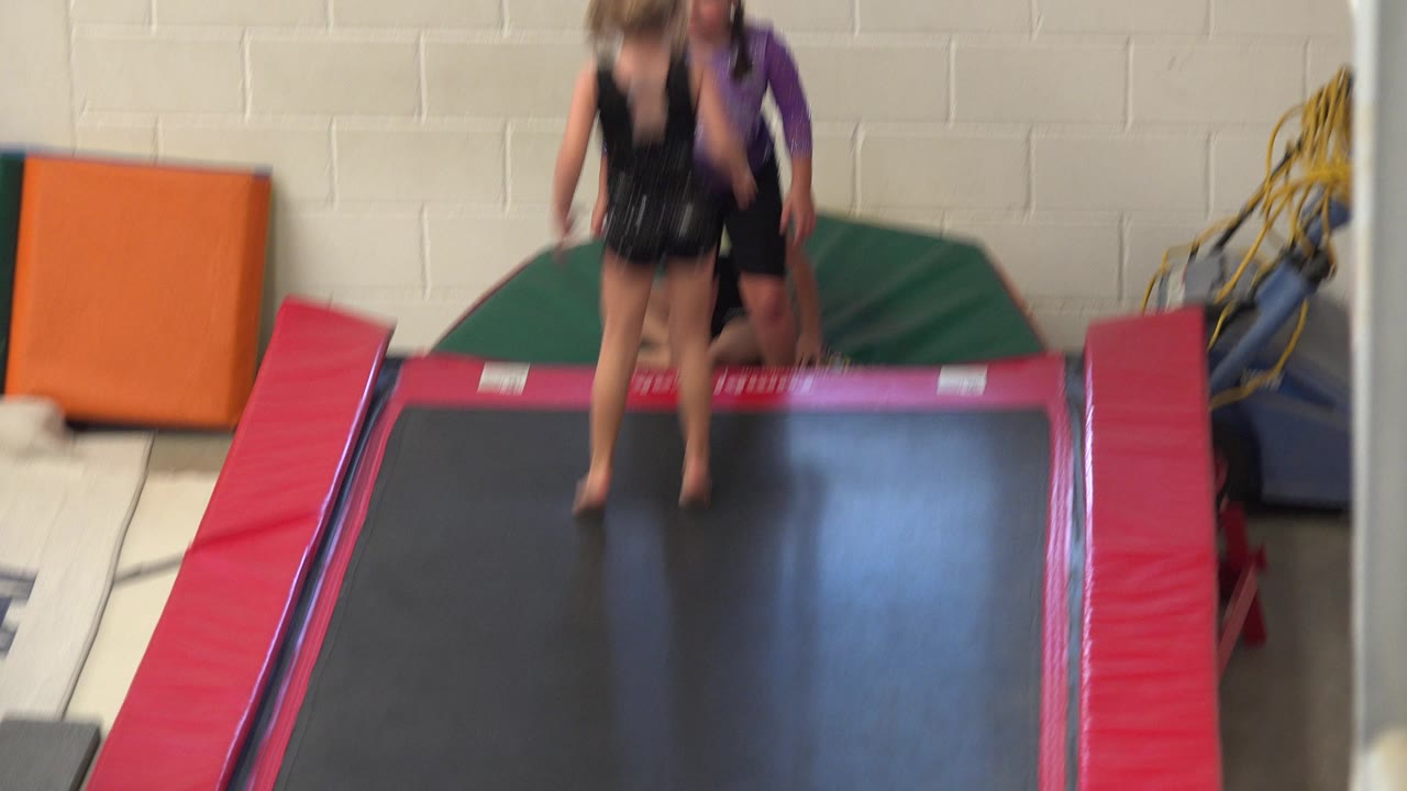 The elevated camera frames two children at the far end of the trampoline in Cabo San Lucas, Mexico, with pink side pads and stacked gym mats visible around them.