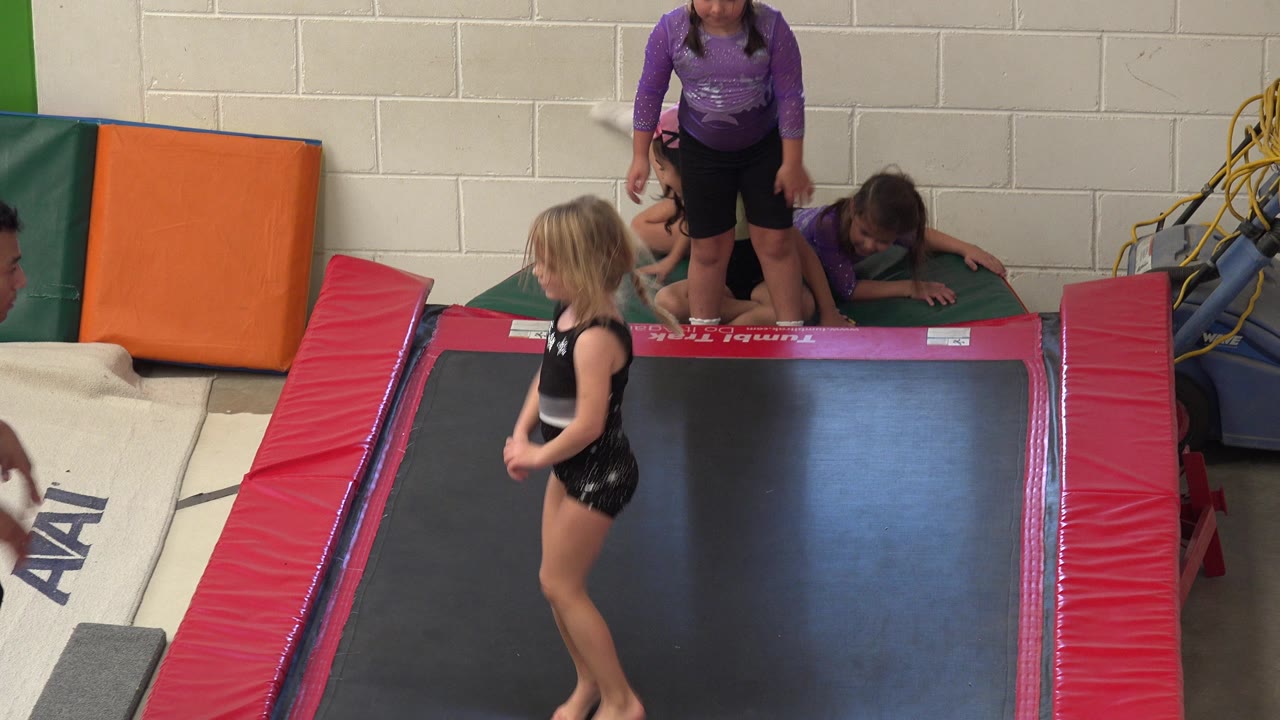 Young girl stands on a trampoline while children wait on mats behind her.
