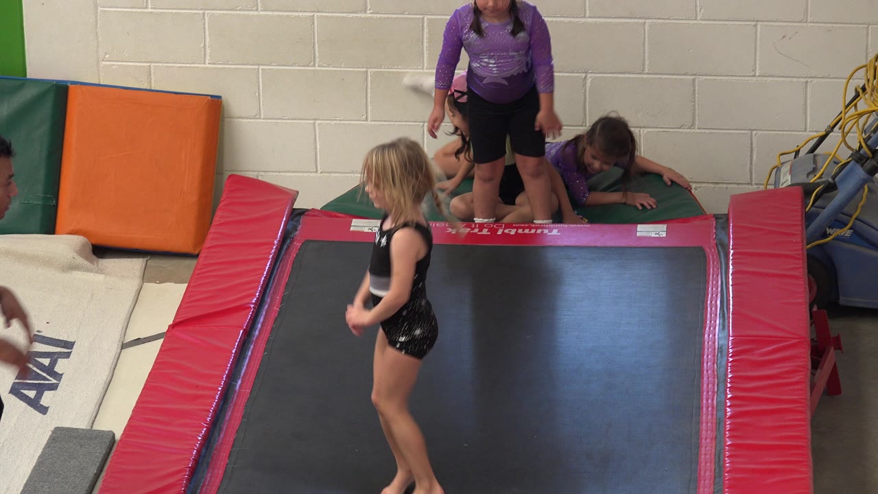 In Cabo San Lucas, the young gymnast remains on the black trampoline while children behind her gather near padded mats and wall equipment inside the gym.