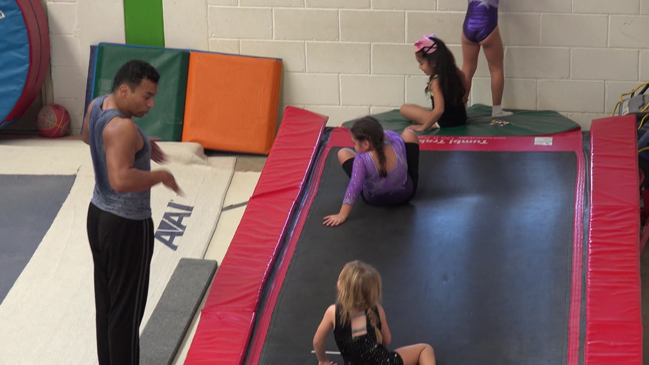 In Cabo San Lucas, Mexico, a male trainer stands beside a black trampoline while two young girls sit on the mat and another child is partly visible on the rear platform.