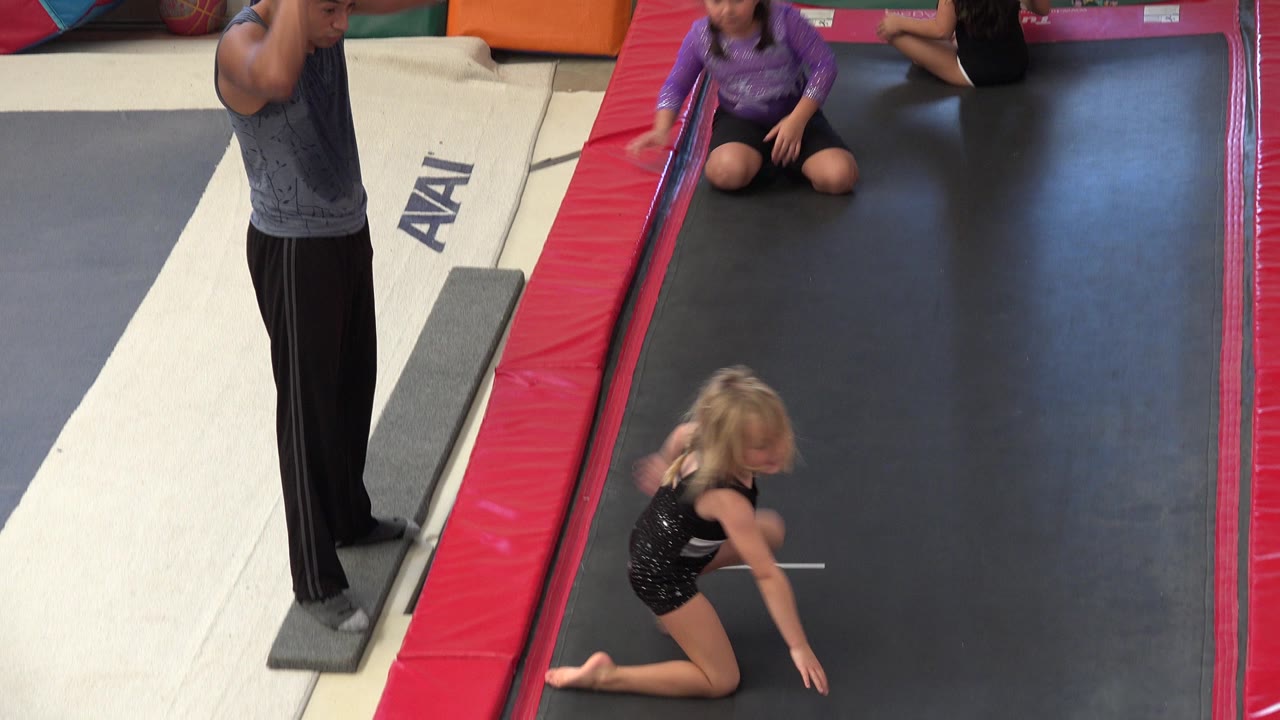 One second in, the blonde child remains low on the black platform as the adult stands at the left edge and another child in purple sits at the far end in the Cabo San Lucas indoor gym scene.