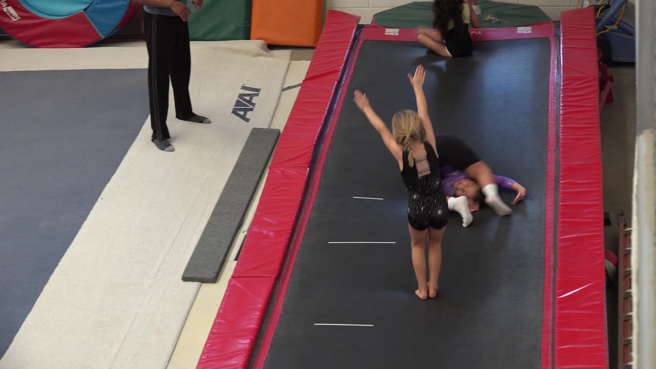 Two young gymnasts on a black trampoline lane with a coach standing nearby.