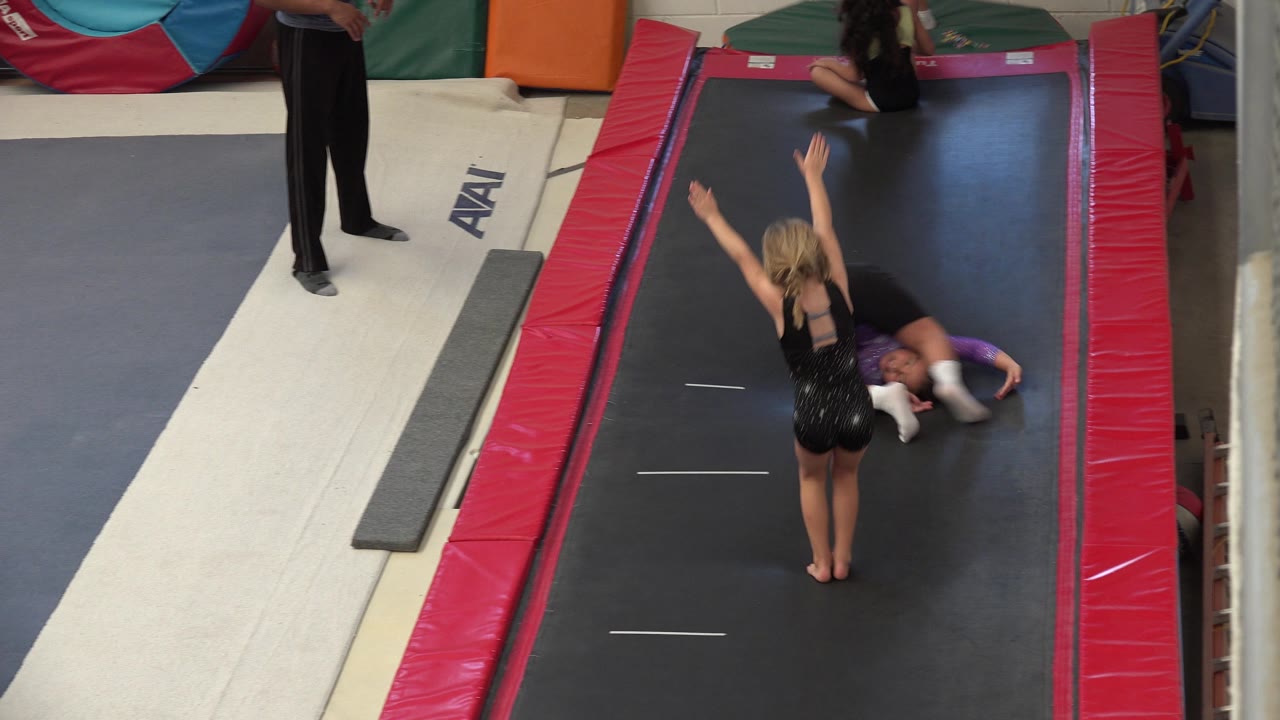 A child in a black leotard raises both arms on the trampoline lane as another child lies near the side and the coach's legs are visible in the Cabo San Lucas gym.