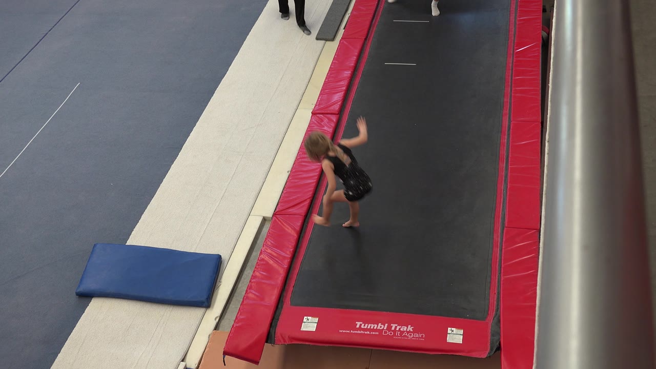 The child gymnast bends near the red trampoline border in Cabo San Lucas, with the blue crash mat and gray floor mats positioned to the left.
