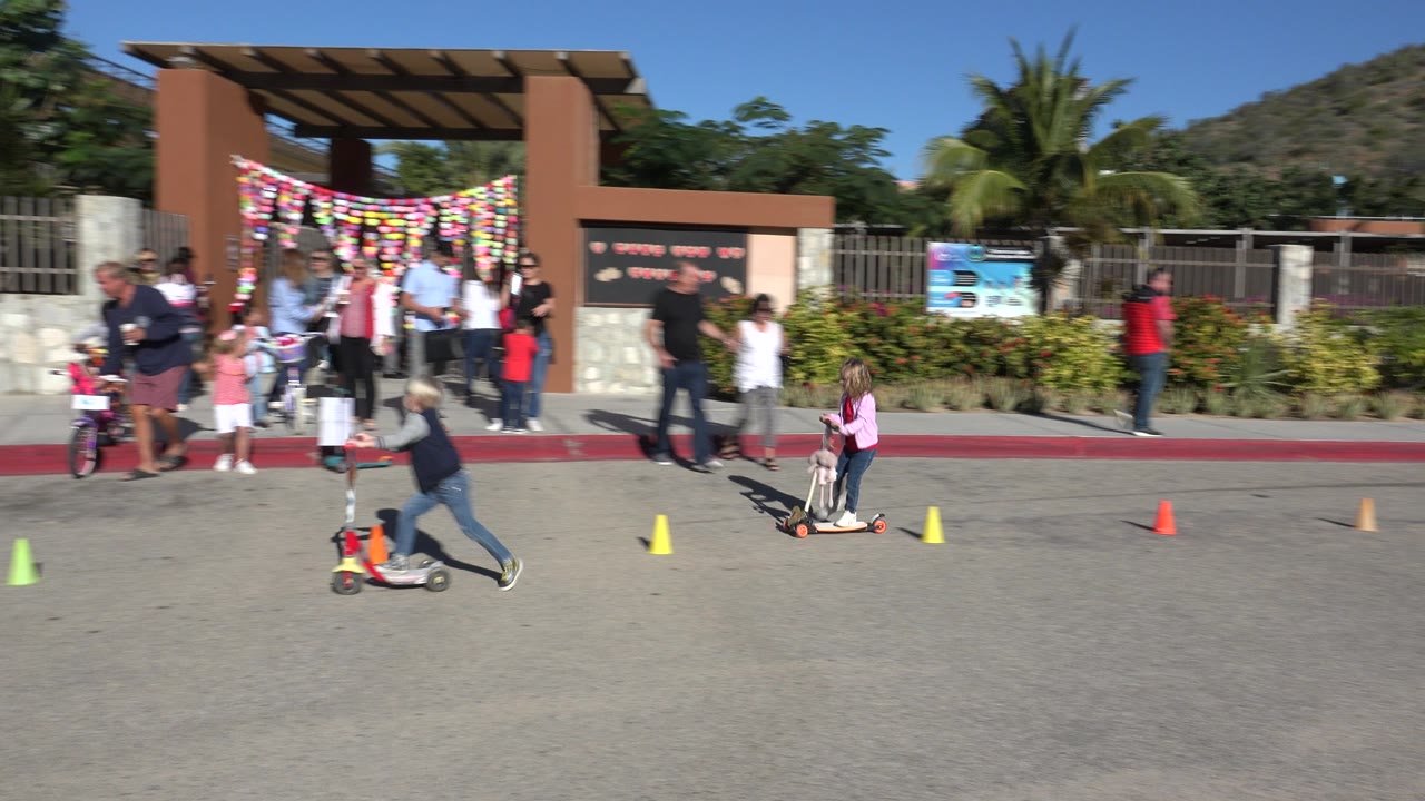 Children ride scooters and a small bike near colored cones outside a decorated entrance in Cabo San Lucas, Mexico, with adults standing behind them.