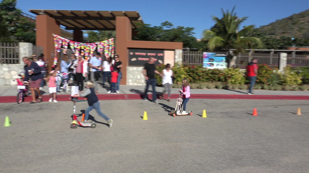 Two children ride scooters across the sunlit street in Cabo San Lucas, Mexico, passing yellow, orange, and green cones while adults stand by the decorated entrance.
