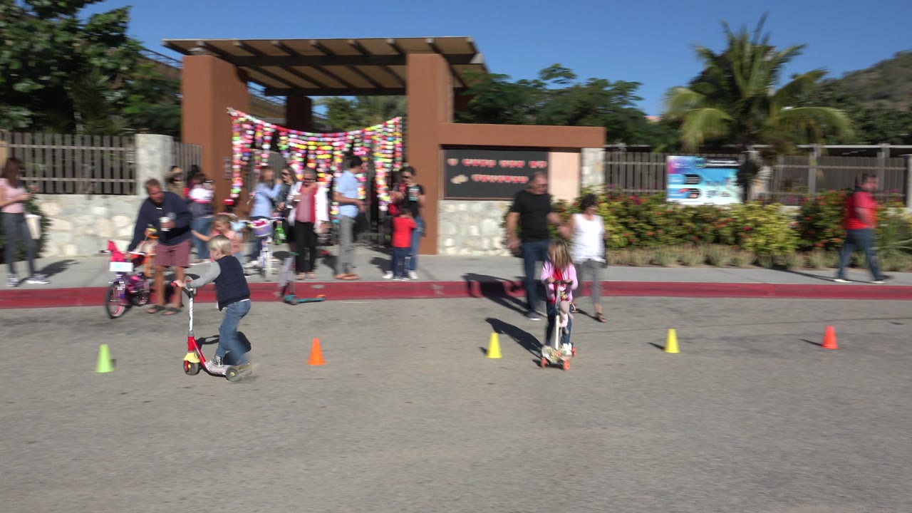 Children ride scooters and a bike near a group of adults.