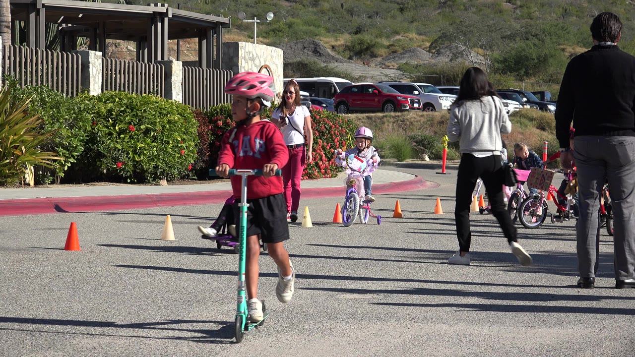 Child in a pink helmet rides a scooter past children on bikes and adults watching.