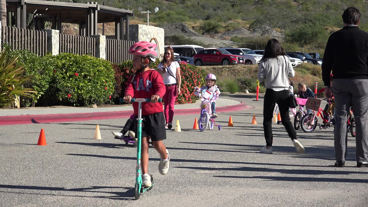 In Cabo San Lucas, the scooter rider moves farther across the foreground while a child on a purple bicycle and other children ride behind adults near orange cones.
