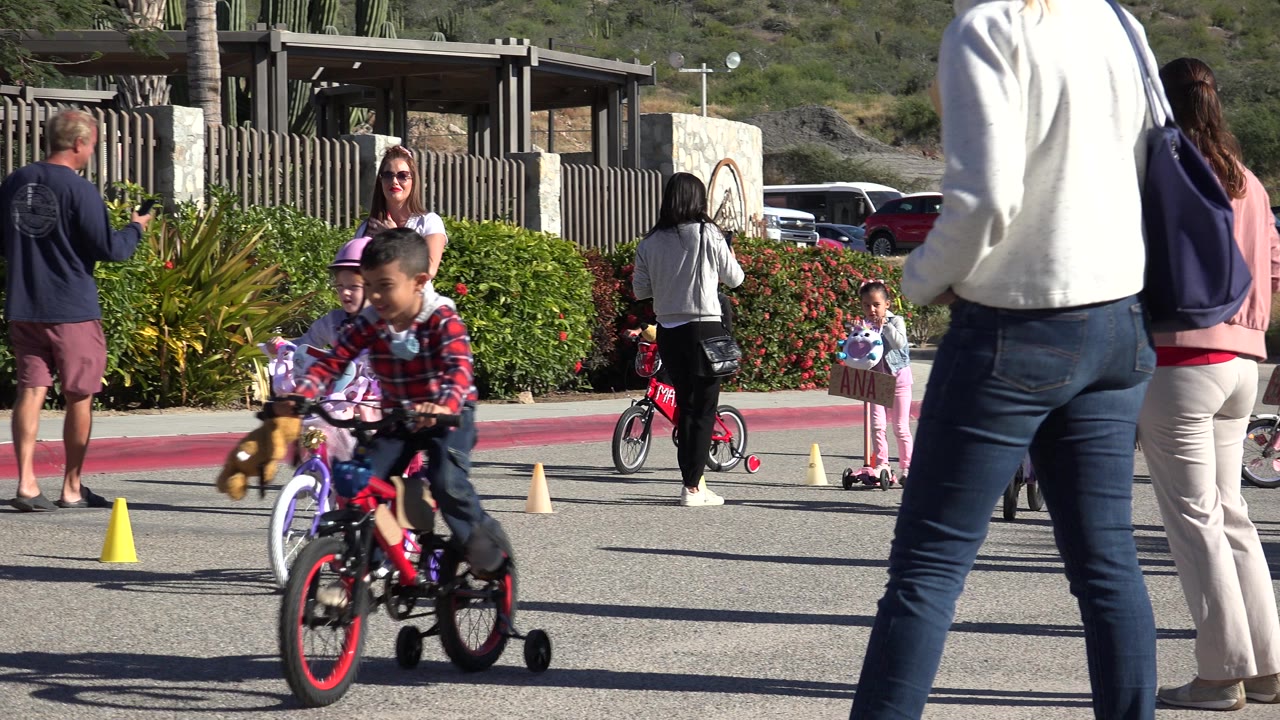 Children practice riding small bicycles around traffic cones in Cabo San Lucas, Mexico, with adults supervising beside shrubs and parked cars.