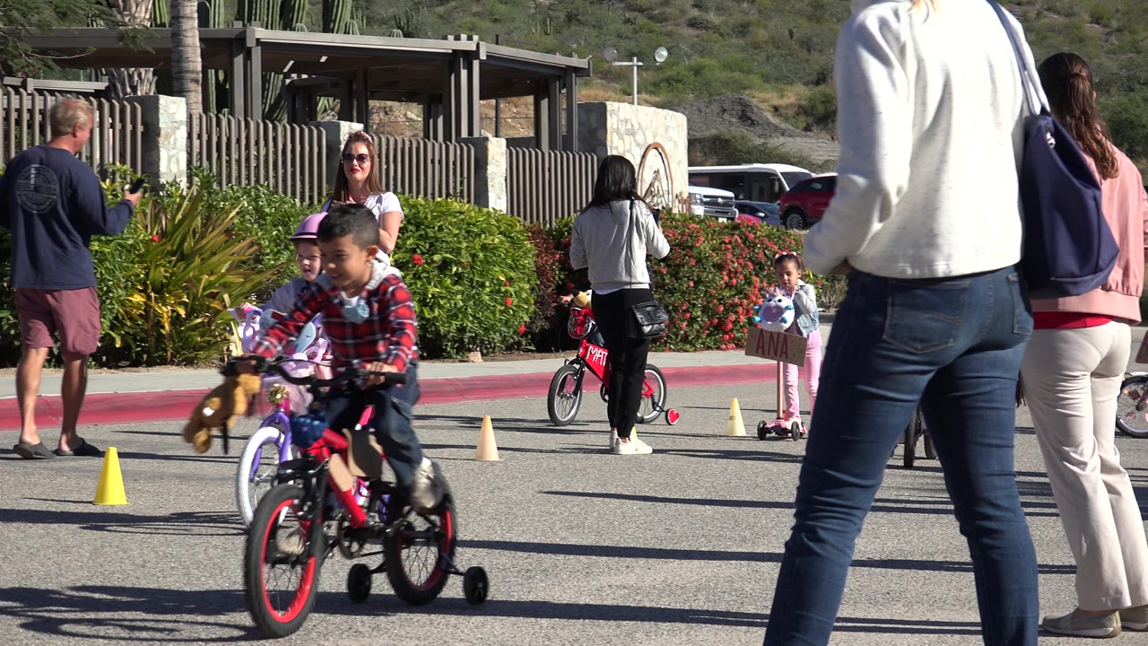 A child on a small bicycle moves through the foreground while other children and adults stand around the cone-marked practice path in Cabo San Lucas, Mexico.