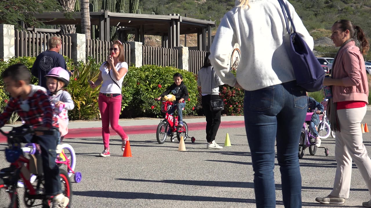 A child on a bike passes close to the camera with adults and cones behind.