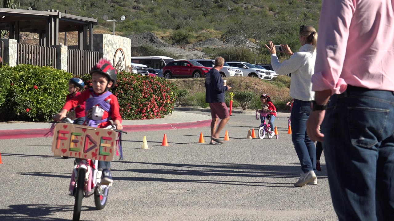 Child riding a small bicycle toward camera near cones and adults.