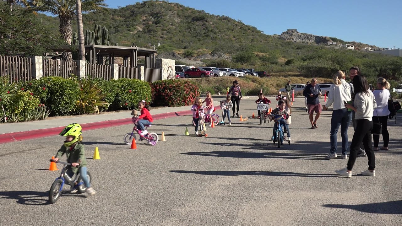 Children ride bicycles and scooters past cones as adults watch on a sunny street.