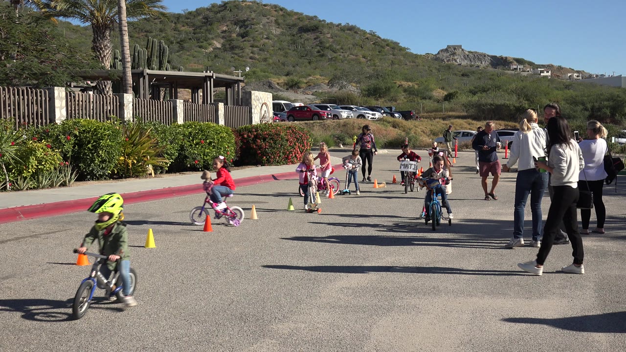 A child wearing a bright helmet rides across the foreground in Cabo San Lucas as other children continue the bike and scooter race behind adults watching from the roadside.