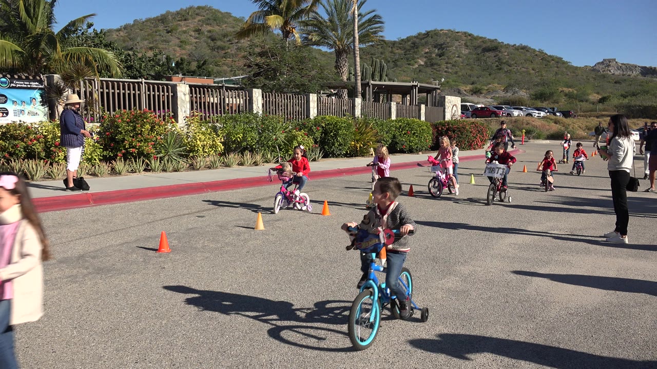 Children ride bicycles and scooters around orange cones on a paved road in Cabo San Lucas, Mexico, with adults, hedges, palms, and hills behind them.