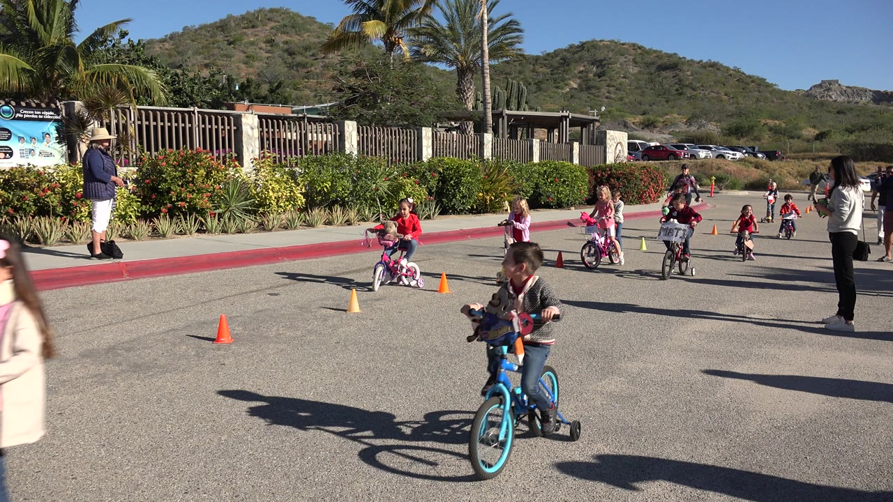 A child on a blue bicycle moves near the foreground as other children ride through cones in Cabo San Lucas, Mexico, with adults watching from the road edge.