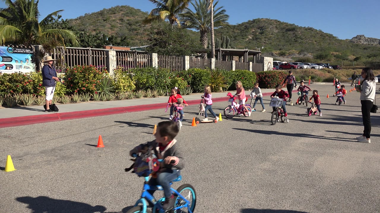 Several children ride bikes and scooters between cones with a woman standing nearby.