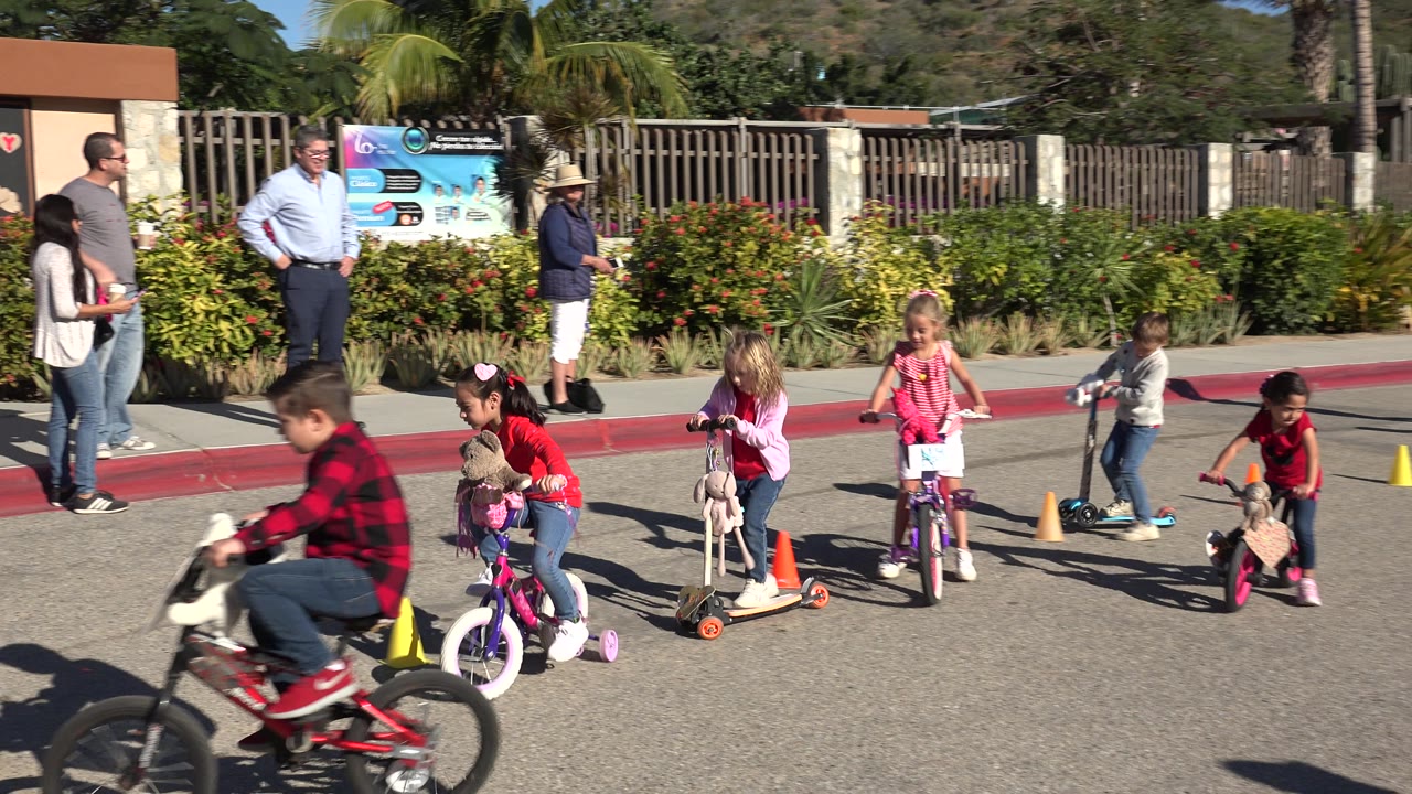 Children riding small bikes and scooters near adults by a park entrance.