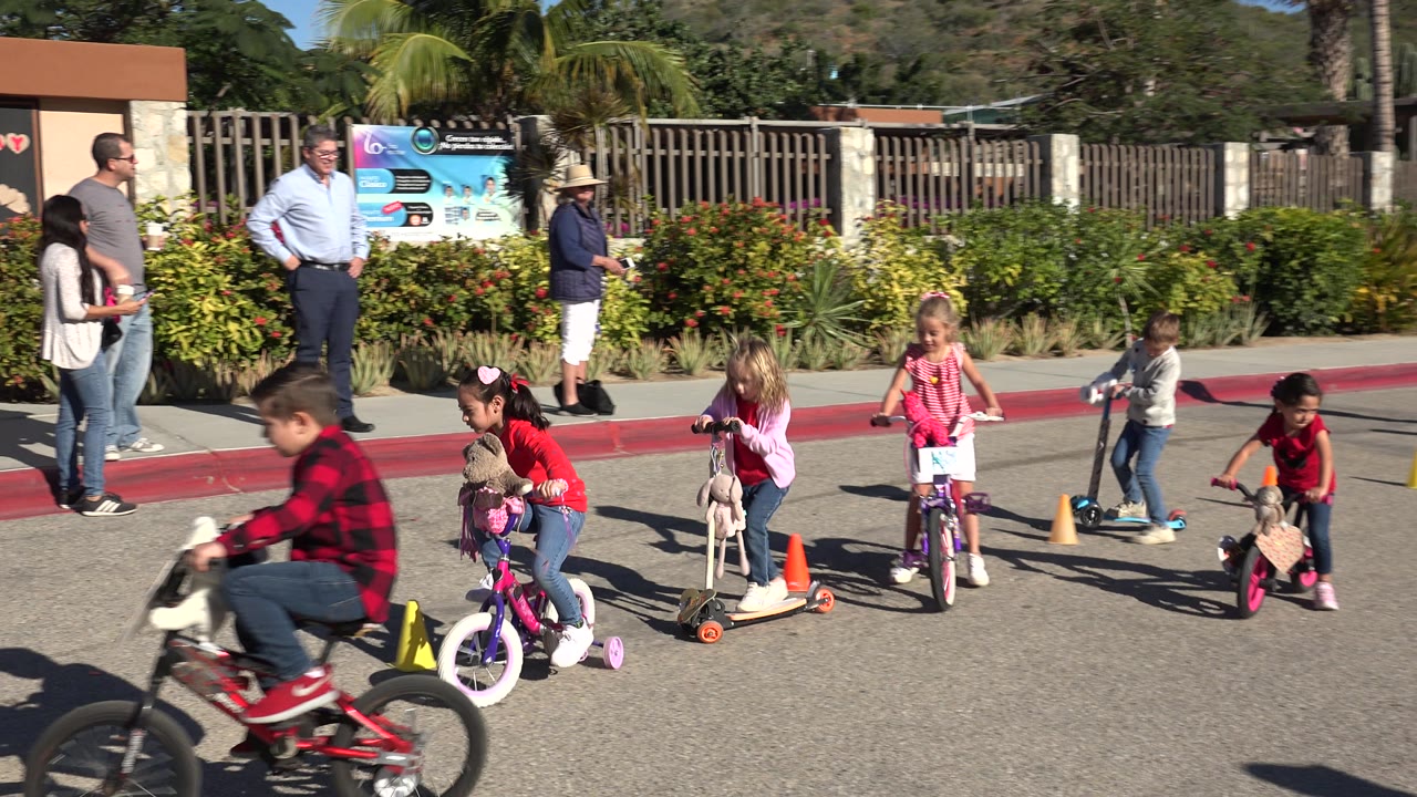 A boy on a bicycle passes close to the camera as children continue riding scooters and bikes in Cabo San Lucas, with adults and colorful signs behind them.