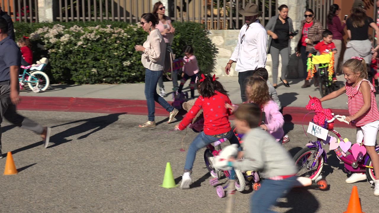 Girls on small bikes and scooters move past colored cones in Cabo San Lucas, with a woman walking at the curb and other adults and children in the background.